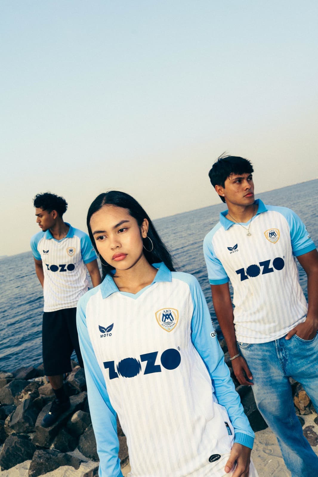 Three people wearing AIO Mytilinis jerseys by the sea